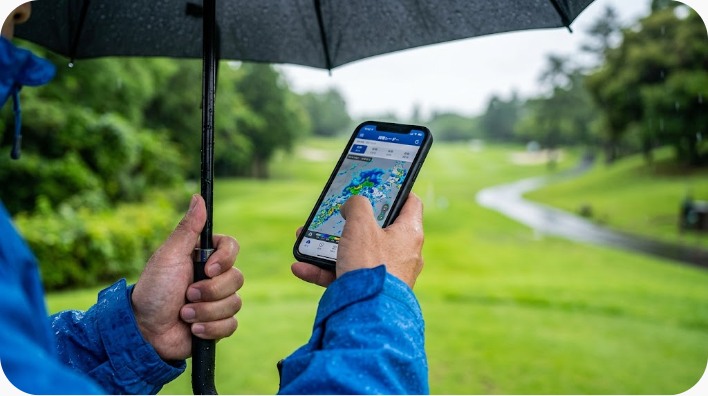 レインウェアを着たゴルファーが傘を差してスマートフォンの雨雲レーダーを確認している手元のクローズアップ