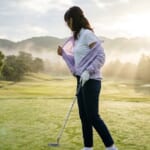 朝霧の残る練習グリーンでウインドブレーカーを脱ぐ女性ゴルファー